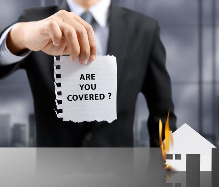 An Executive holding a piece of paper stating ARE YOU COVERED? Beside, house made out of paper and burning 