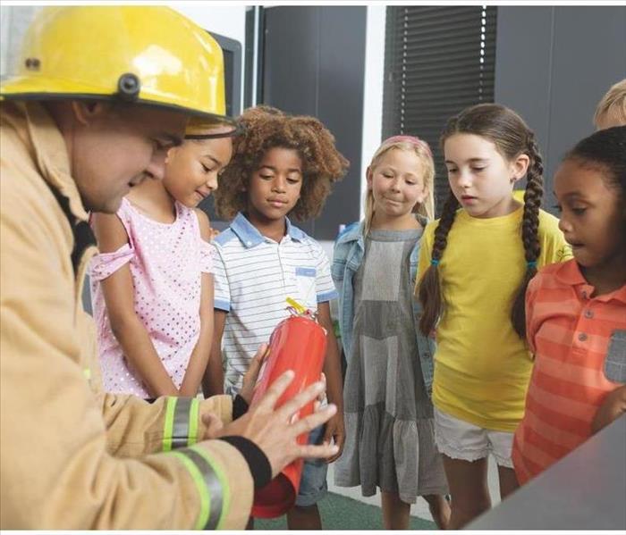 Rear view of a Caucasian firefighter wearing a fire helmet over his head and teaching about fire extinguisher to school kids 